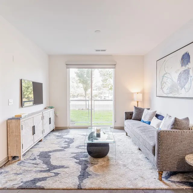 Contemporary Living Room Design A modern living room featuring a gray sofa with decorative cushions, a glass coffee table, and a light-colored area rug. A large window provides natural light and an outdoor view. A TV is mounted on the wall, and an art piece is displayed above the sofa. There is a lamp and a console table in the room.