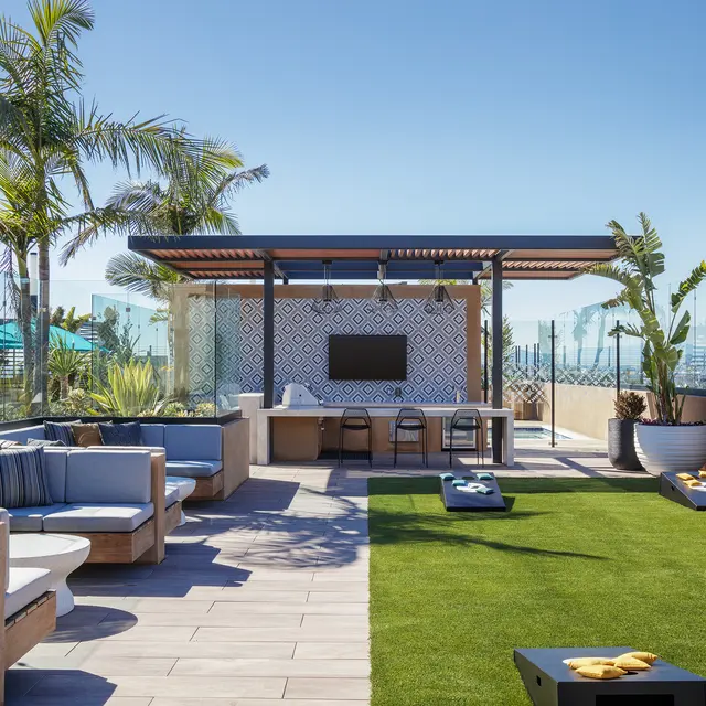 A modern rooftop lounge area featuring comfortable seating, a bar area, and a grass surface. The space is surrounded by glass panels and has tropical plants, with a clear blue sky in the background.