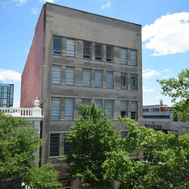 A mid-century building with a plain façade, featuring multiple windows, surrounded by trees and a street view. The sky is clear with a few clouds.