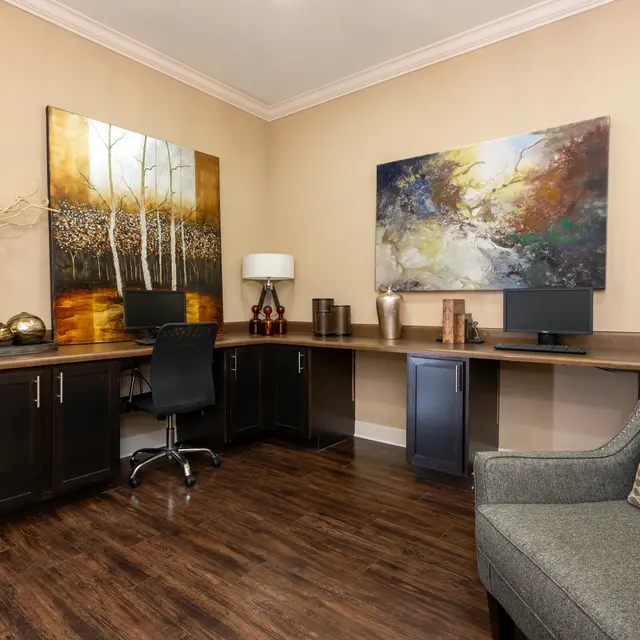 A modern home office featuring a large wooden desk, a black swivel chair, and two artworks on the wall. There are decorative vases and a lamp on the desk, with a cozy gray armchair in the foreground.