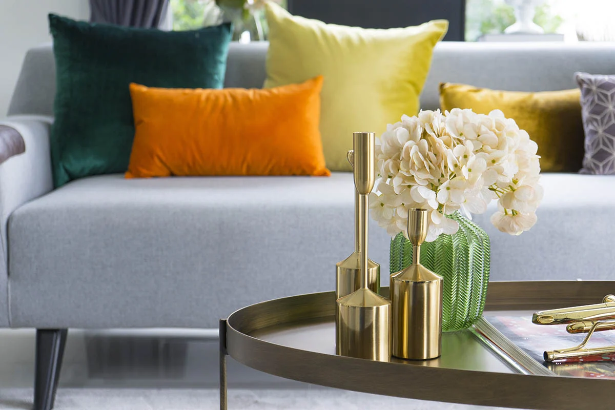 Cozy Modern Living Room Decor A cozy living room featuring a light gray sofa adorned with colorful cushions in green, orange, and yellow. A stylish coffee table holds two gold candlesticks and a green vase filled with white flowers, set against a backdrop of a bright window.