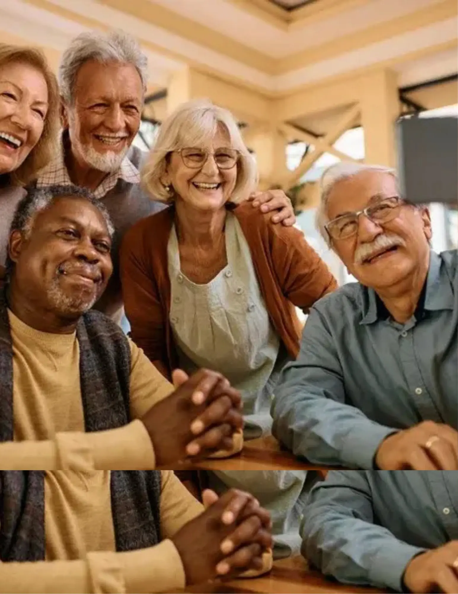 A group of five elderly individuals smiling and posing for the camera in a bright indoor setting. They are sitting around a table, with joyful expressions. One man is in the foreground with his hands clasped, while others are behind him showing enthusiasm and happiness.