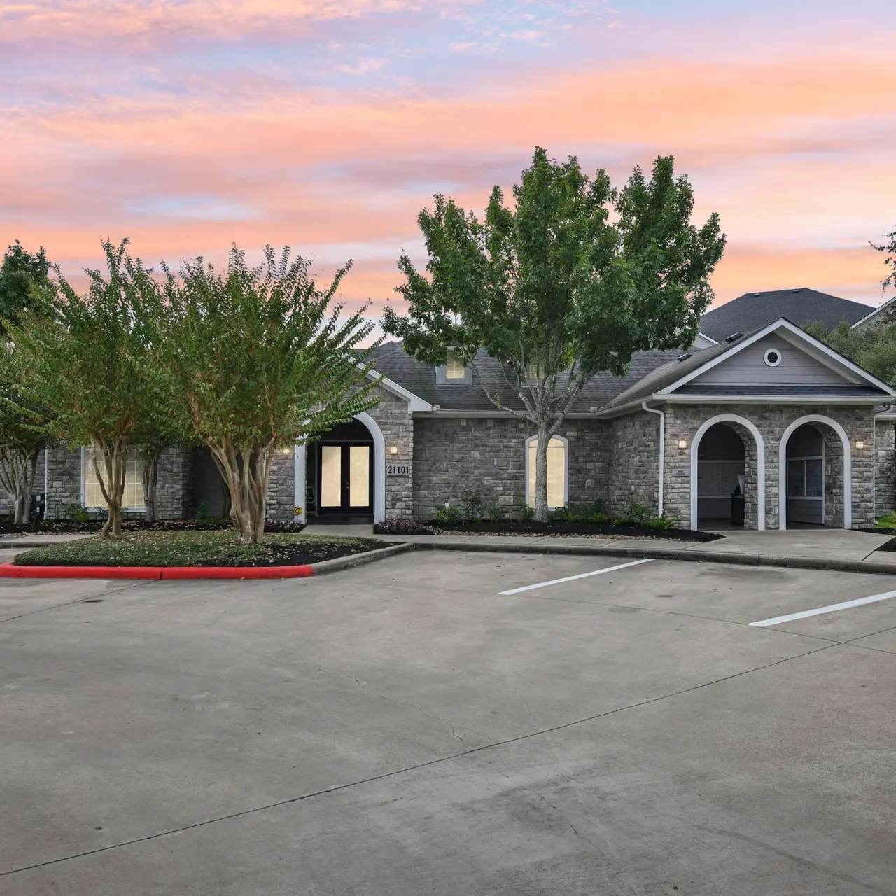 Exterior view of an apartment complex during sunset, featuring a stone entrance and surrounding greenery.