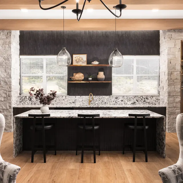A modern kitchen featuring a large island with a marble countertop. There are two black bar stools at the island, and above it hang two pendant lights. The kitchen has stone walls, wooden cabinetry, and decorative shelves displaying various items. Large windows allow natural light to enter, illuminating the space. There are two patterned armchairs in the foreground.