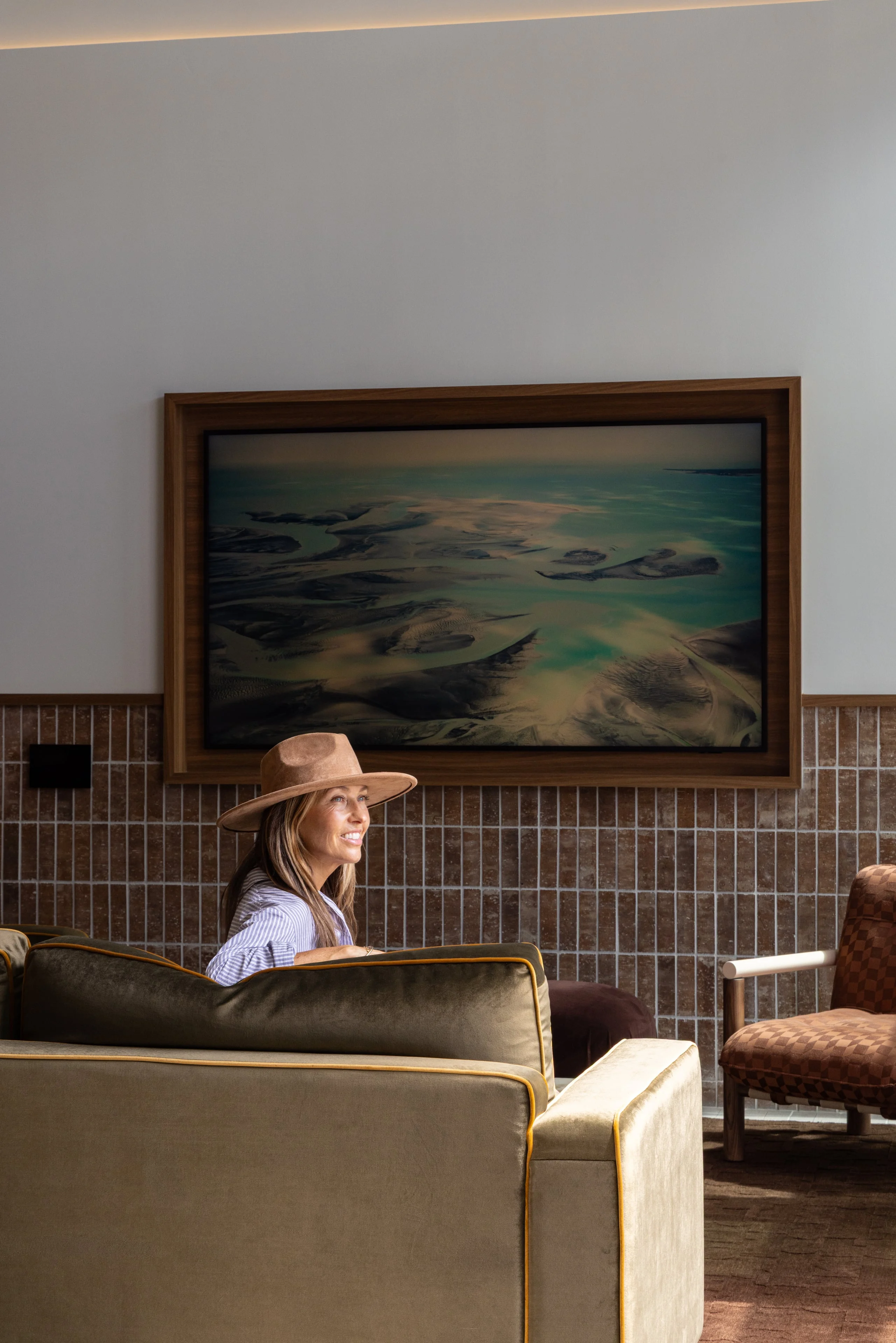 A woman with long hair and a wide-brimmed hat sitting on a couch, looking back towards the camera. Behind her is a large framed painting depicting abstract waves, with a brown tiled wall beneath.