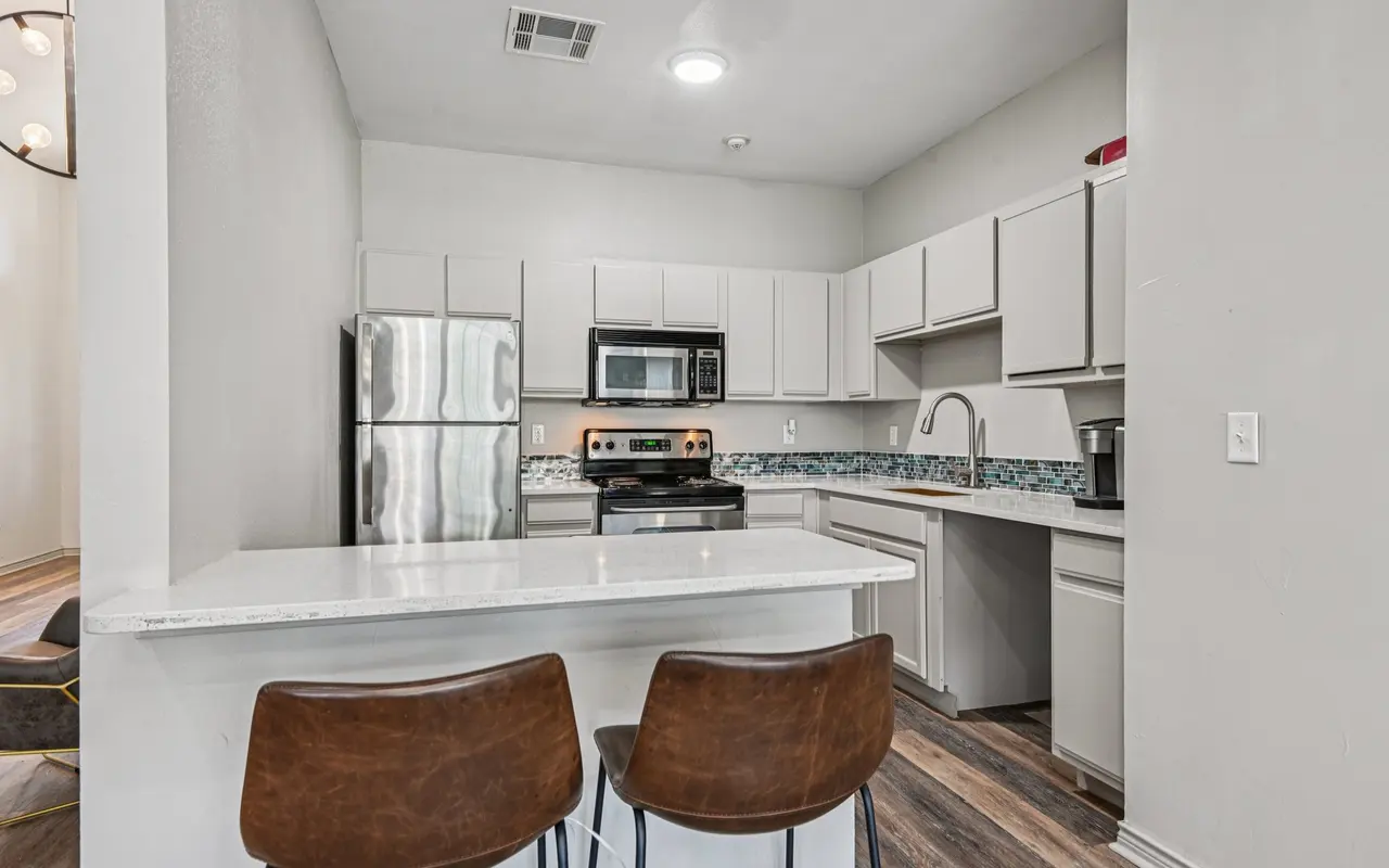A modern kitchen featuring stainless steel appliances, grey cabinets, and a white countertop with two brown bar stools at the counter.