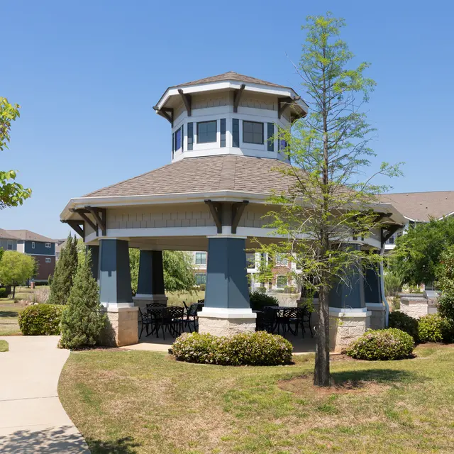 A pavilion-style gazebo surrounded by greenery in a park-like setting, with a clear blue sky above.