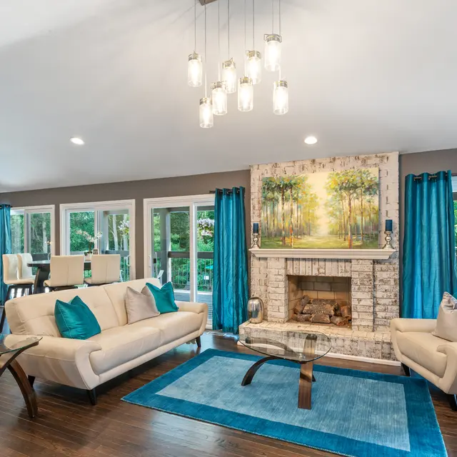 A modern living room featuring beige couches with teal cushions, a glass coffee table, a built-in fireplace, and large windows with teal curtains.