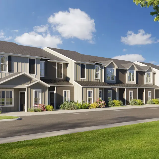 A row of modern townhouses with well-maintained landscaping and a paved road in front. The buildings feature a combination of light-colored siding and darker roofs, with large windows allowing ample natural light.