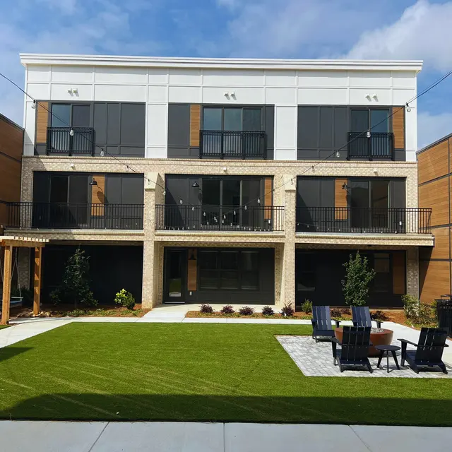Modern Apartment Complex with Green Courtyard A modern apartment complex featuring two levels, with a grassy courtyard in front and lounge chairs. The building has a blend of wood and stone exterior.