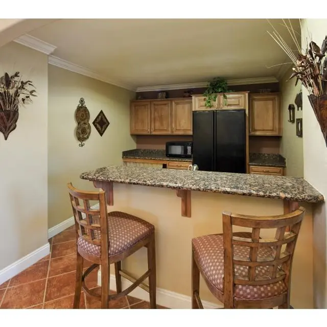 Cozy Kitchen Bar Area A small kitchen bar area with two wooden stools, a granite countertop, and wooden cabinetry. A black refrigerator and microwave are visible in the background, along with decorative wall hangings and neutral paint colors.