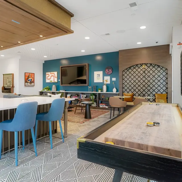 An interior view of a modern lounge and game area featuring a long table, blue chairs, a wall-mounted television, and decorative artwork.