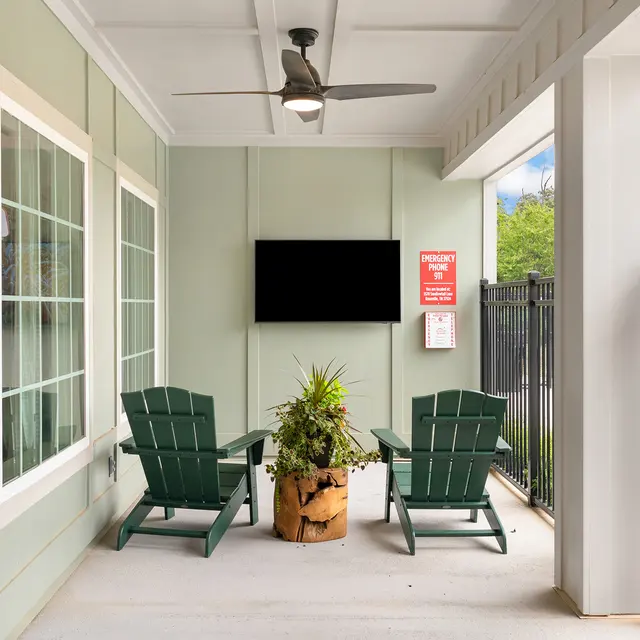 An outdoor lounge area featuring two dark green Adirondack chairs facing a small, potted plant and a flat-screen TV mounted on the wall.