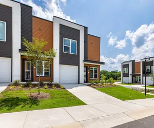 A row of modern two-story townhouses with colorful facades, grassy lawns, and concrete driveways under a bright blue sky with fluffy clouds.