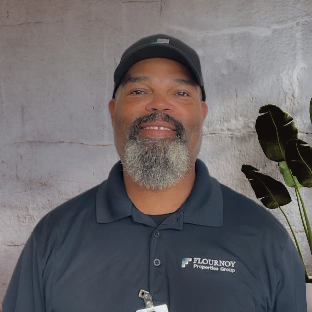 A smiling man with a beard, wearing a black shirt with a logo, stands in front of a textured, light-colored background. He is sporting a cap and appears friendly and approachable.