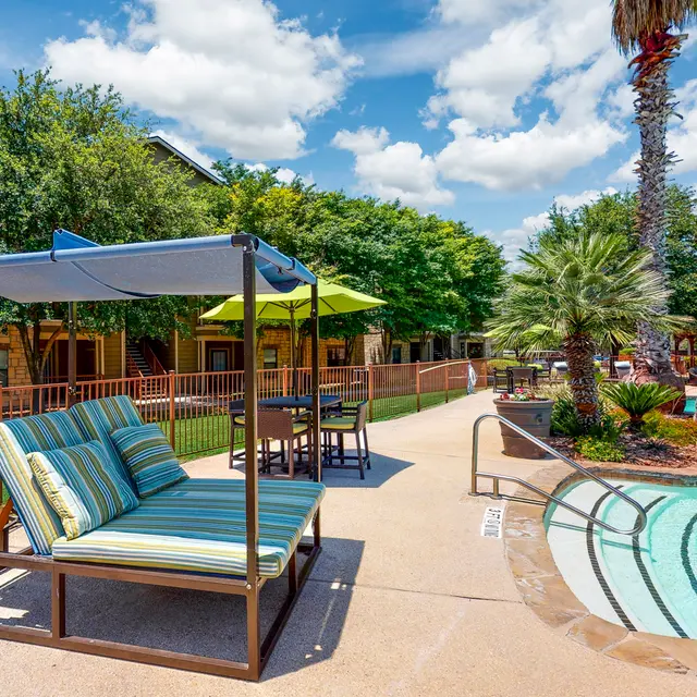 Relaxing Poolside Lounge Area A scenic view of a poolside lounge area featuring a striped lounge chair, a shade canopy, and patio tables under green umbrellas, surrounded by lush trees and palm plants on a sunny day.