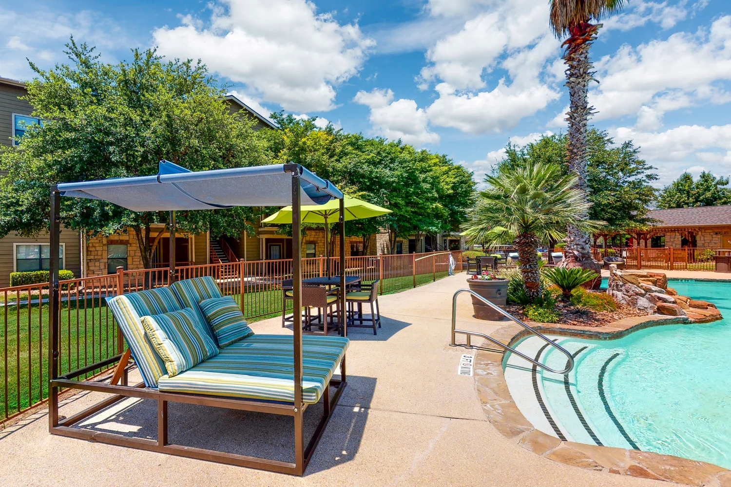 Relaxing Poolside Lounge Area A scenic view of a poolside lounge area featuring a striped lounge chair, a shade canopy, and patio tables under green umbrellas, surrounded by lush trees and palm plants on a sunny day.