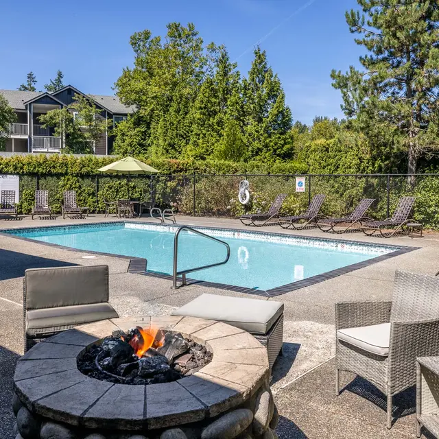 A tranquil poolside area featuring a turquoise swimming pool surrounded by lounge chairs and green umbrellas. There is a stone fire pit in the foreground with a small flame, and lush greenery in the background.