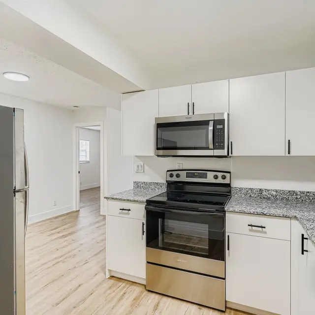 A modern kitchen featuring a stainless steel refrigerator, an electric oven, and a microwave. The kitchen has white cabinets with black handles and a granite countertop. The flooring is light-colored wood and there is a doorway leading to another room in the background.