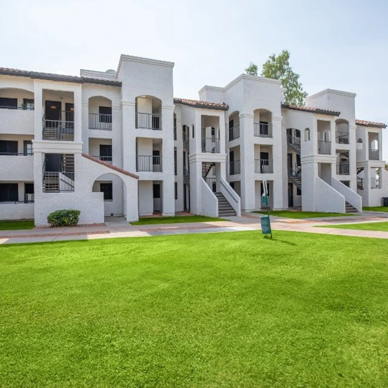 A multi-story apartment complex with white stucco walls and green lawns in front. The building features several staircases and balconies.