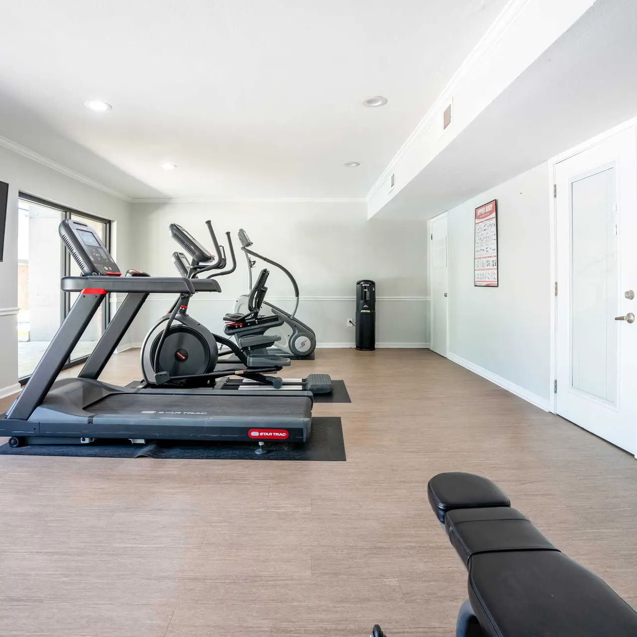 A well-lit gym featuring a treadmill, elliptical machine, and a bench, with a television mounted on the wall and large windows letting in natural light.