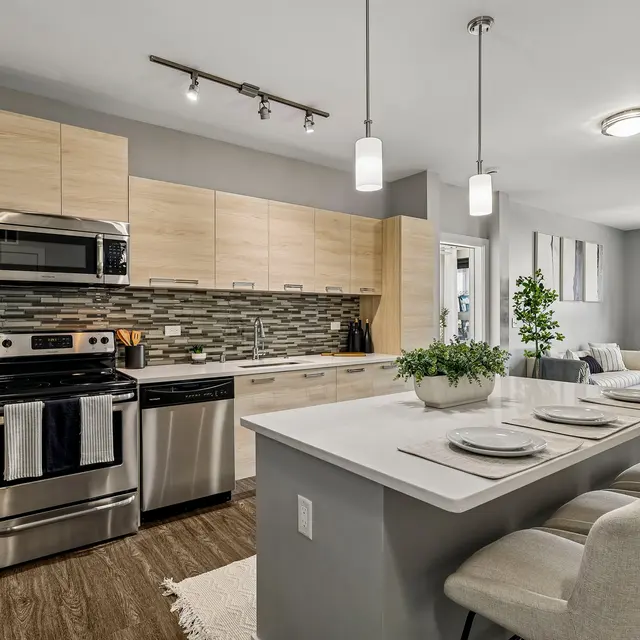 A modern kitchen featuring light wood cabinetry, stainless steel appliances, and a gray tile backsplash. The kitchen island includes seating and is adorned with a small plant. Natural light illuminates the space through large windows.