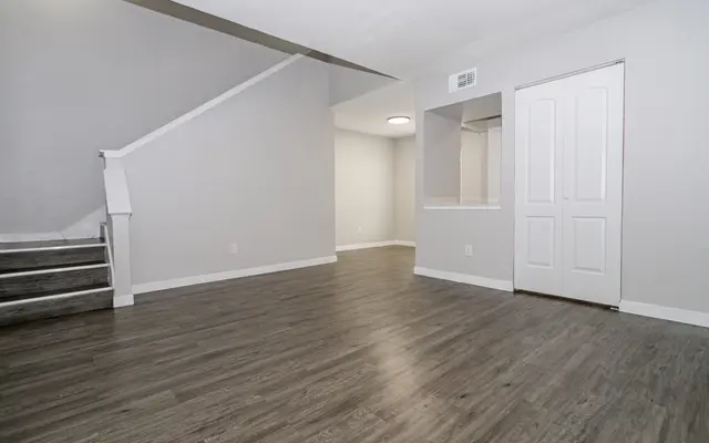 A spacious, empty living room with gray walls, wood-like flooring, and a staircase on the left. There's a doorway on the right leading to another room.
