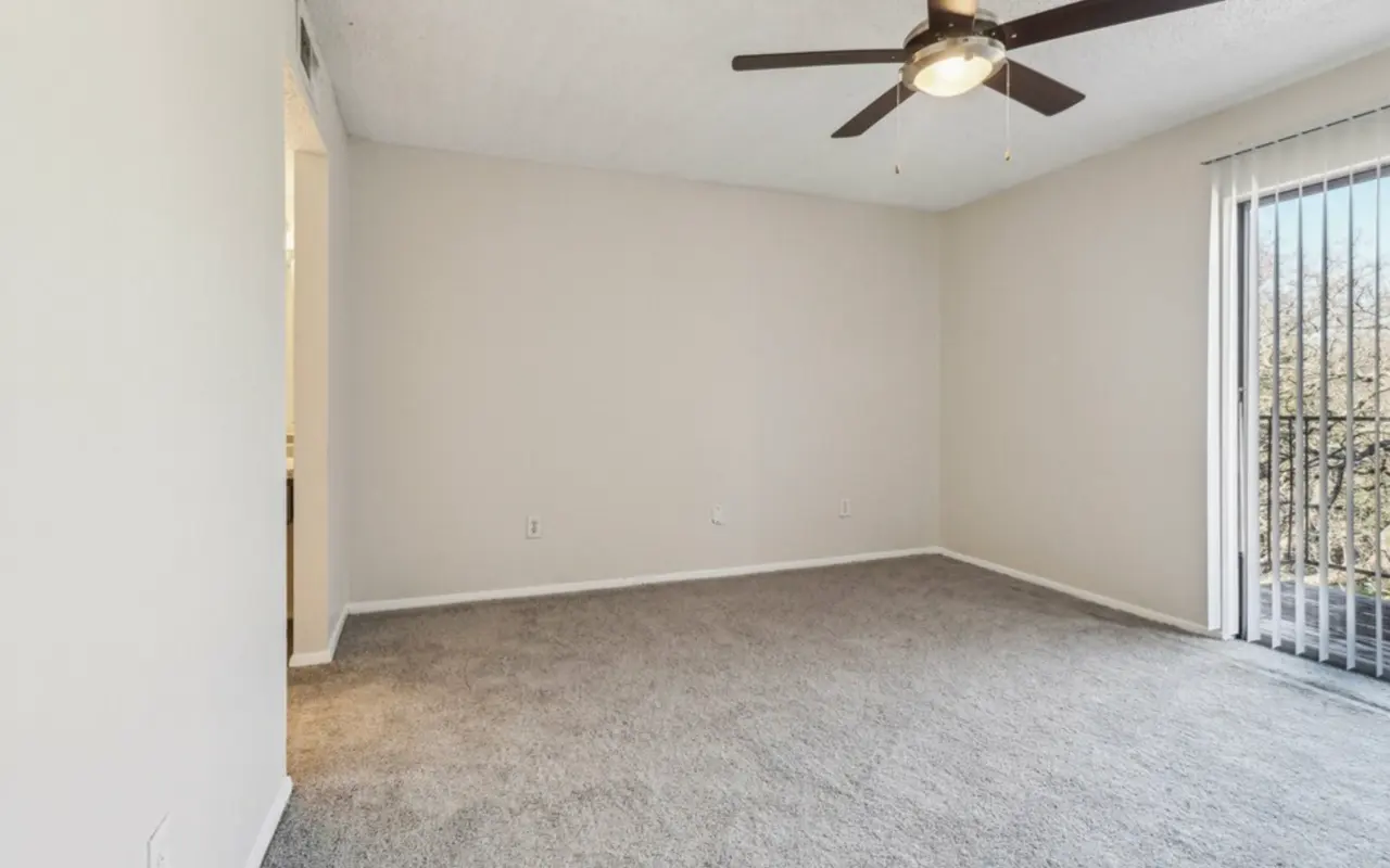 A spacious, empty living room with light-colored walls, a ceiling fan, and large sliding glass doors leading to a balcony. The room has soft carpet flooring and minimal furnishings.
