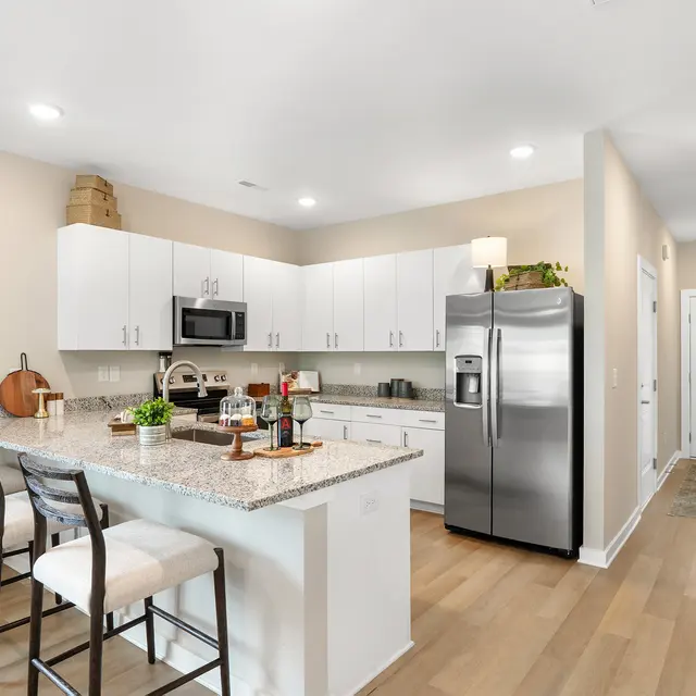 A modern kitchen featuring white cabinets, stainless steel appliances, and a granite countertop with seating. A decorative pumpkin and plants are on the counter, with warm lighting overhead.