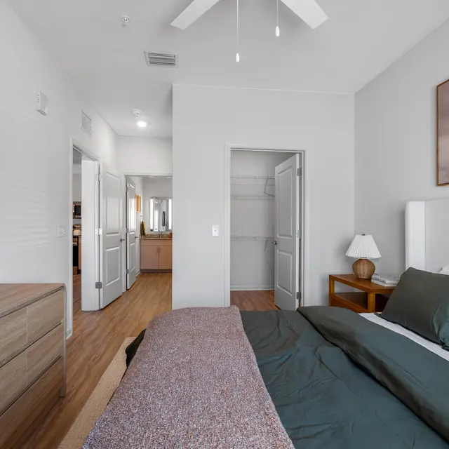 A modern bedroom featuring a neatly made bed with dark green bedding and a blanket draped over it. There is a wooden dresser with a decorative plant on top, a bedside lamp, and artwork hanging on the wall. Two doorways lead to other parts of the space, with one showing a closet.