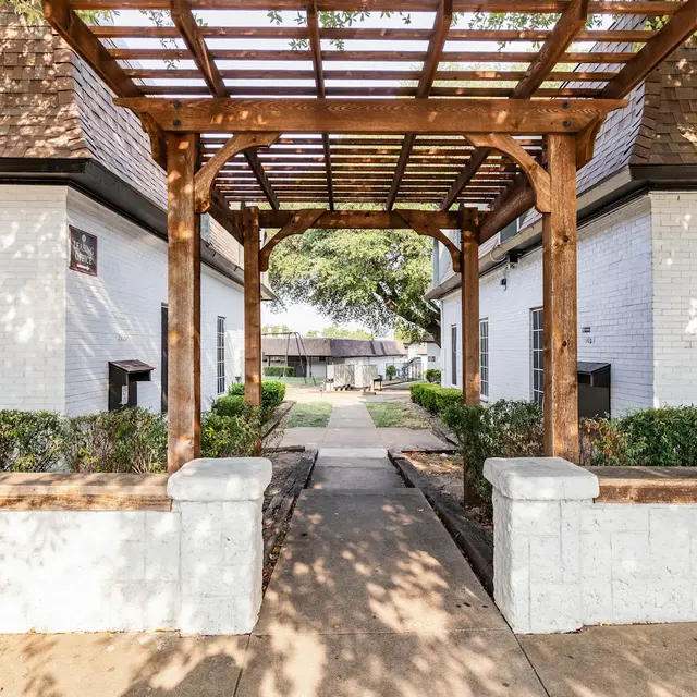 A wide pathway leading through a garden area, framed by a wooden arch. The path is bordered by low green shrubs and the buildings on either side are white with dark roofs. The sky is clear, suggesting a sunny day.