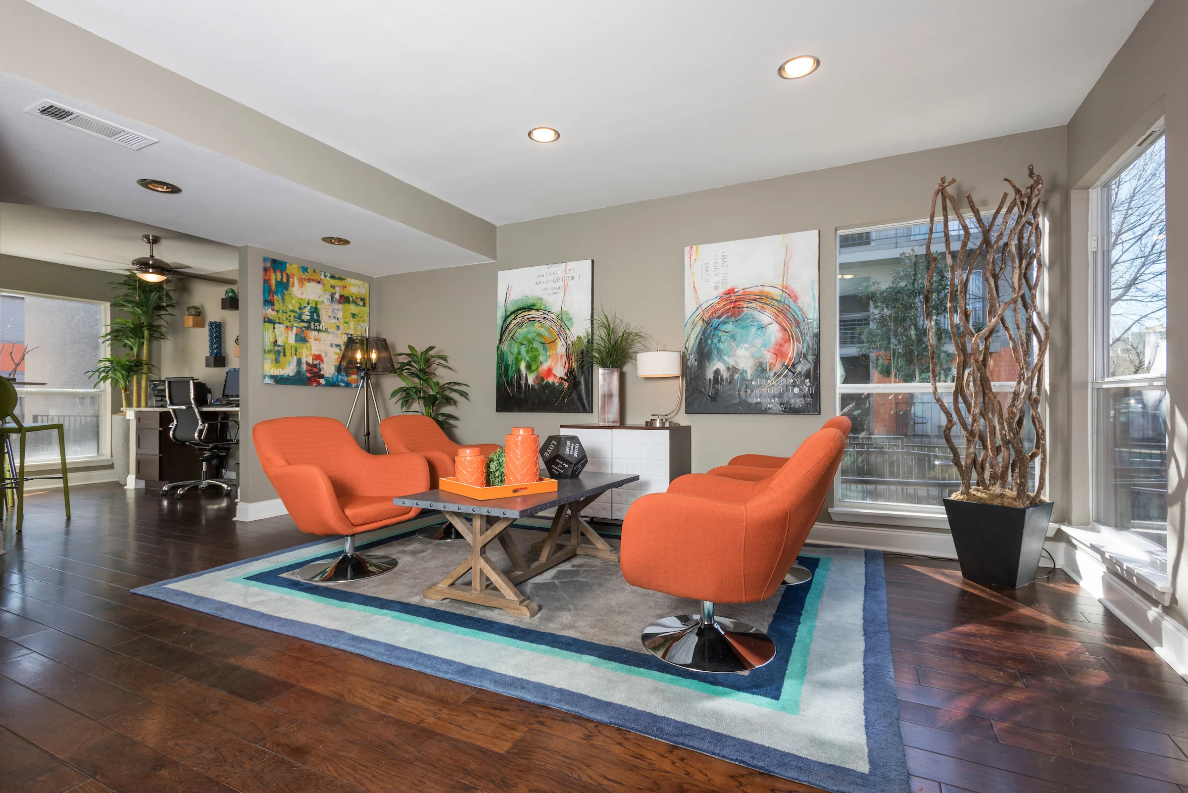 A modern living room with two orange chairs, a wooden table, abstract art on the walls, and large windows letting in natural light.