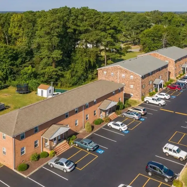 Aerial view of a brick apartment complex with green trees in the background and a parking lot filled with cars.