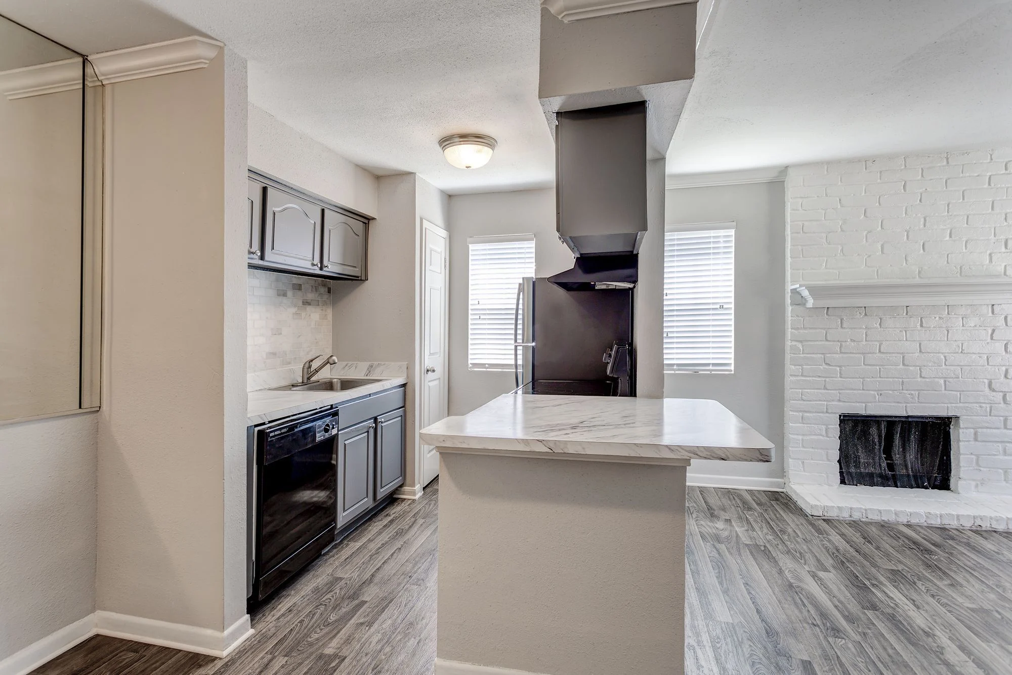 Contemporary Kitchen and Living Space Modern kitchen and living area with gray cabinets, a marble countertop, a black dishwasher, and a brick fireplace.