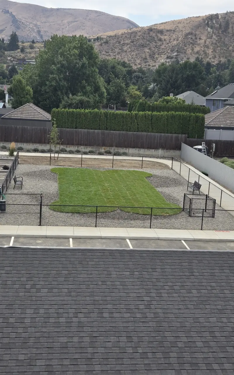 An aerial view of a fenced dog park featuring a grassy area shaped like a bone, surrounded by gravel and a parking area nearby.