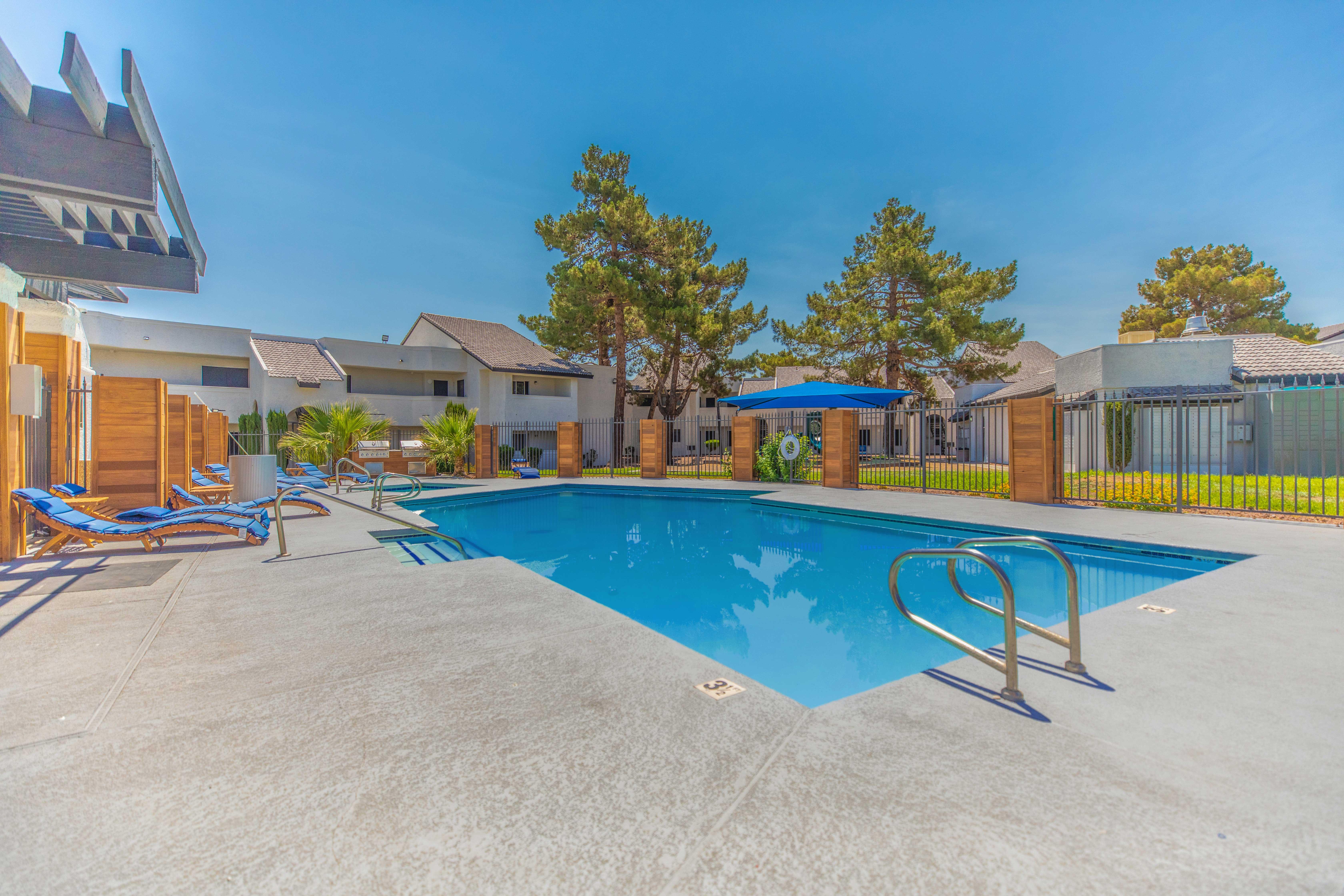 A well-maintained outdoor swimming pool area surrounded by lounge chairs and trees under a clear blue sky.