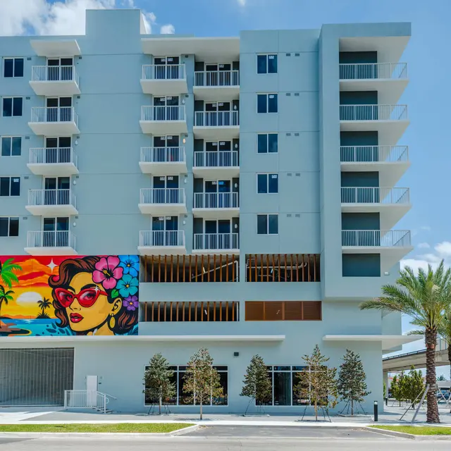 A modern multi-story building with large balconies and a colorful mural on one side, depicting a woman with sunglasses and flowers in her hair, surrounded by palm trees and a clear blue sky.