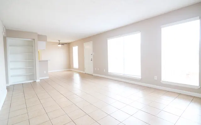 A spacious, empty living room featuring light-colored walls and tile flooring. Large windows allow natural light to fill the space, and a ceiling fan is visible. There is an open area with a small partition leading to another space with shelves.