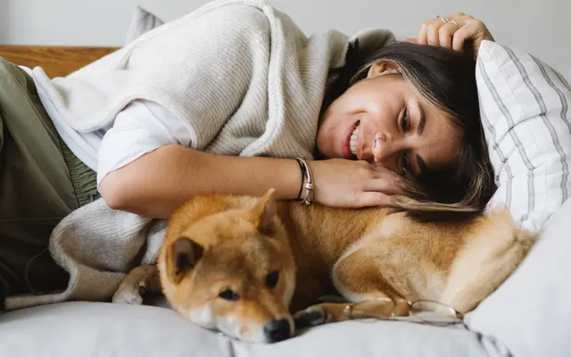 Cozy Home Moment with a Shiba Inu A woman lying on a couch, smiling while resting her head on a Shiba Inu dog. The dog is lying beside her, with a relaxed expression. The setting is cozy, featuring light-colored pillows and warm tones.