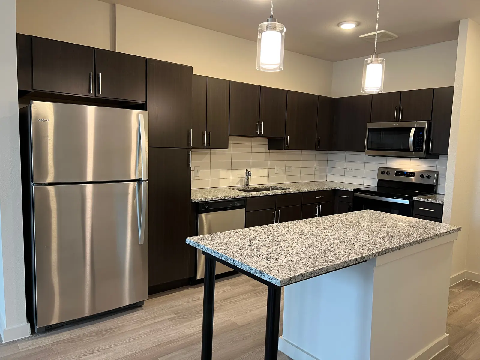 Modern Kitchen Interior A modern kitchen featuring dark wooden cabinets, stainless steel appliances, and a large granite island with bar seating. The kitchen has a light-colored tiled backsplash and warm-toned flooring.