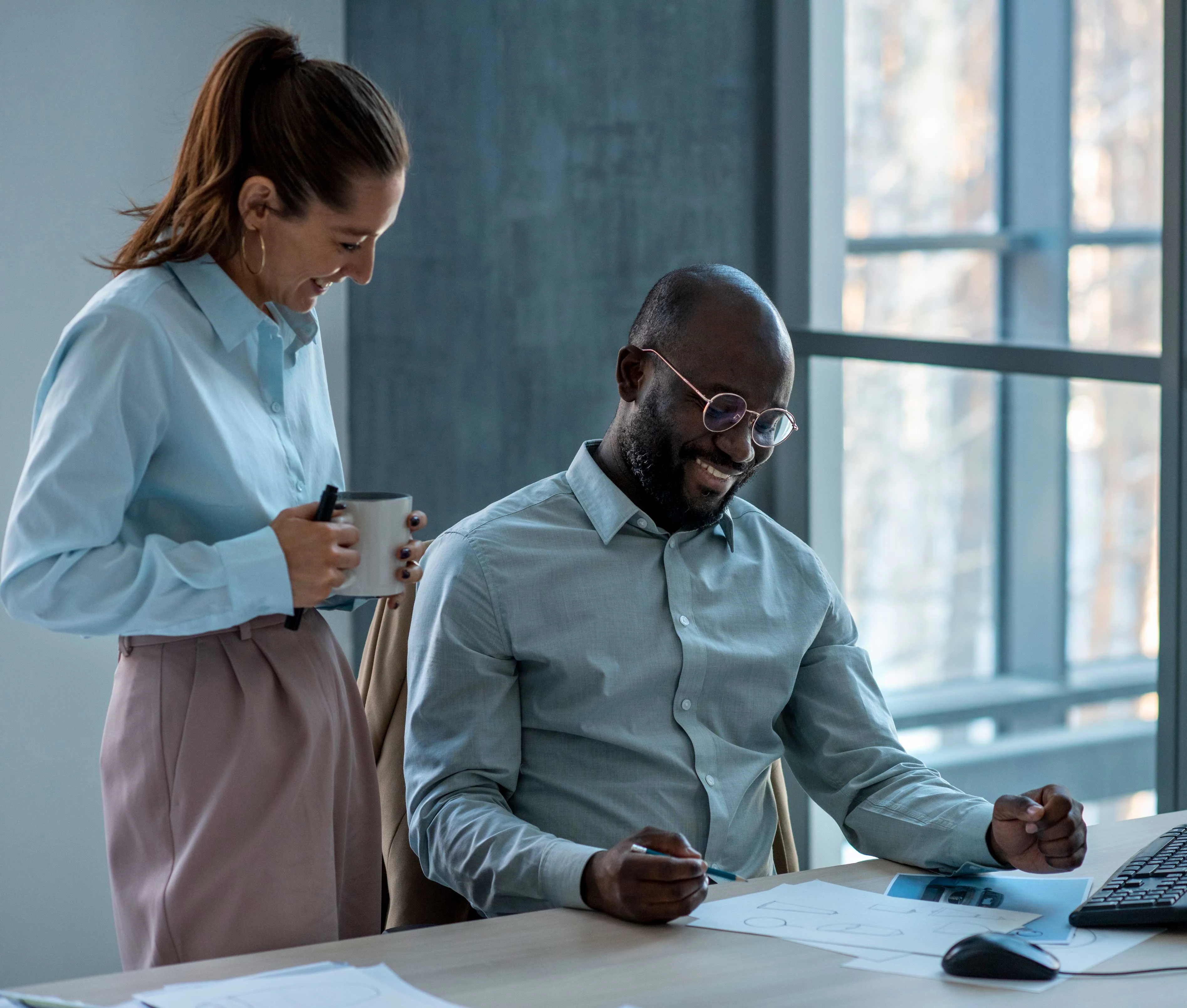 Two colleagues smiling and interacting in an office setting, one holding a cup and the other reviewing documents, with a computer on the desk and large windows in the background.