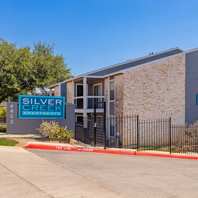 Exterior view of Silver Creek Apartments with a blue sign, surrounded by greenery and a fence.