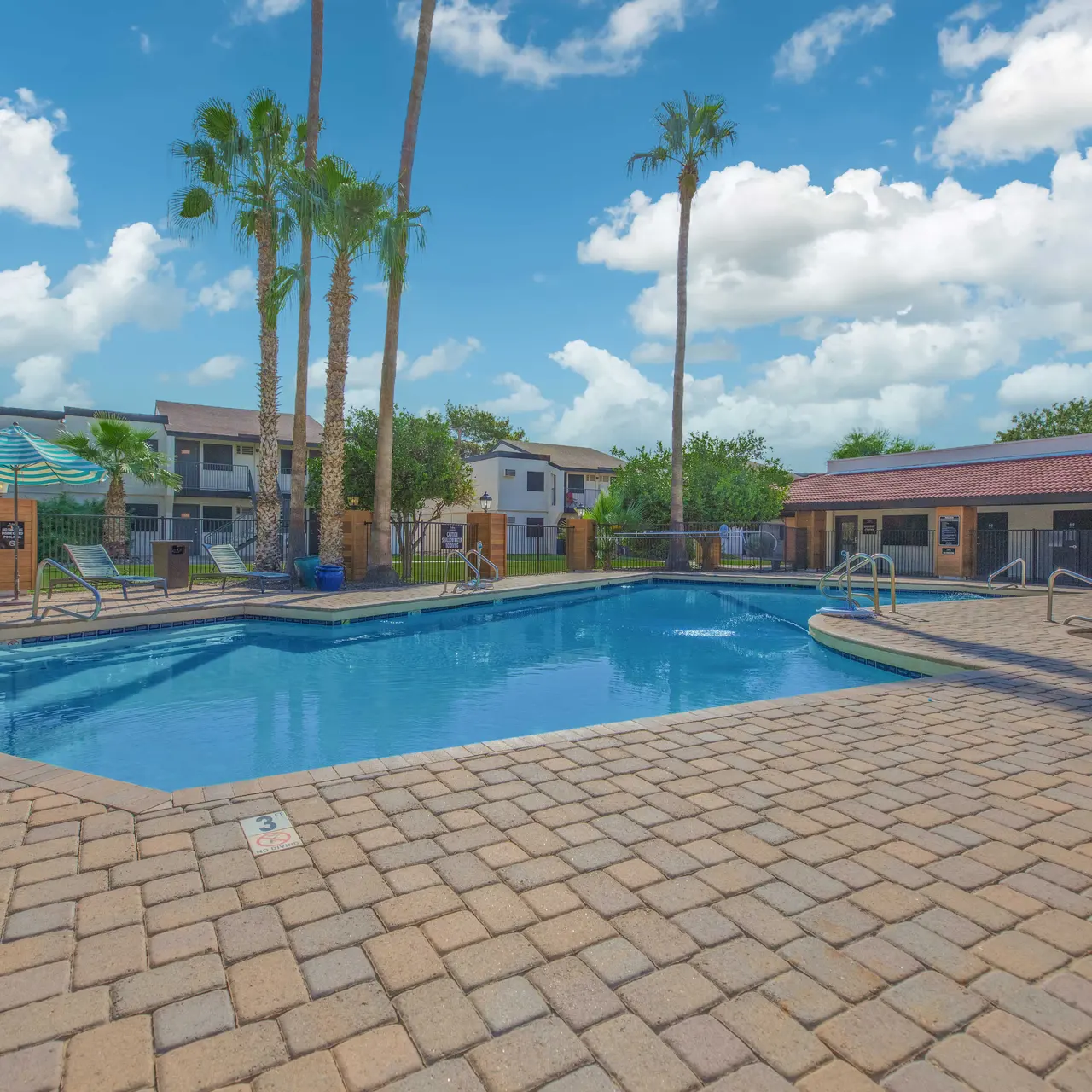 A sunny swimming pool area surrounded by palm trees and apartment buildings, featuring lounge chairs and umbrellas near the pool.