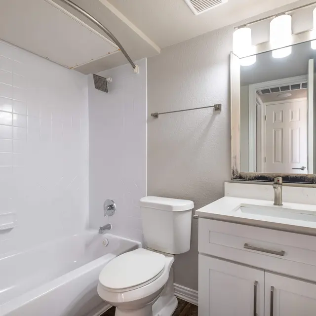 A modern bathroom featuring a white bathtub, toilet, and sink with a mirror above the sink. The walls are light-colored with a tiled bath area.
