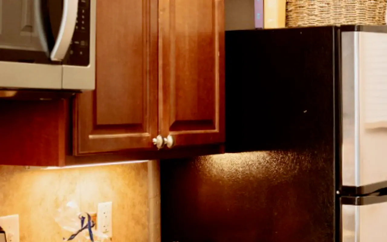 Cozy Kitchen Corner A cozy kitchen corner featuring wooden cabinets, a microwave, and a dark refrigerator. A marble countertop holds a bowl and kitchen towels.