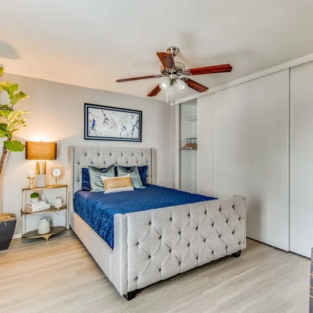 A modern bedroom featuring a tufted bed with a blue bedspread, a bedside lamp on a small table, a large closet, a potted tree, and a decorative dresser.