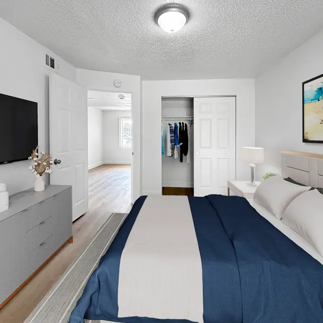 A modern bedroom featuring a king-sized bed with white and navy bedding, a light-colored dresser, a wall-mounted television, and a bright window in the background. A closet is visible through an open door.