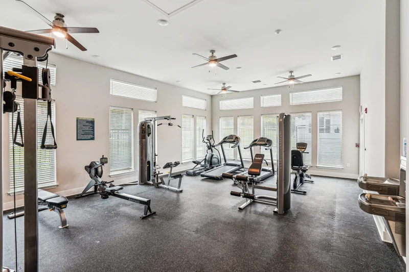 A bright, spacious gym with various exercise equipment, including treadmills, weights, and multiple windows for natural light.
