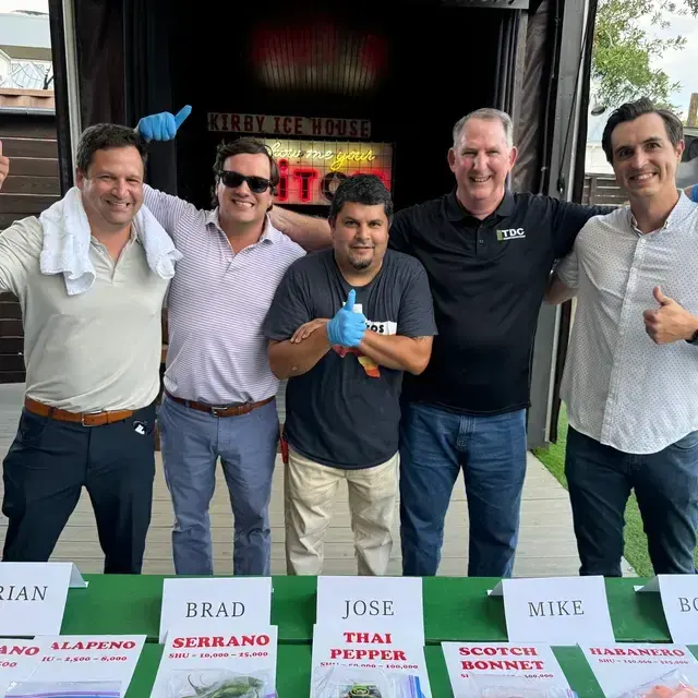 A group of five men standing in front of a sign at Kirby Ice House, each giving a thumbs up. They are dressed casually and smiling, with a table in front displaying names and food items.