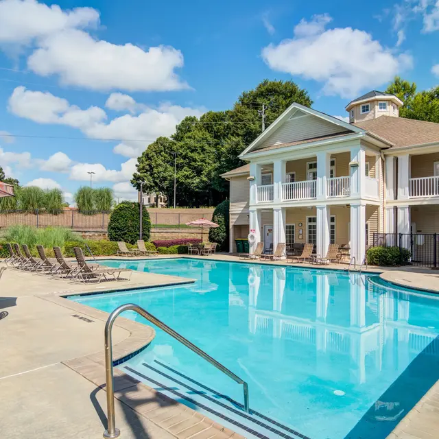 A sparkling blue pool surrounded by lounge chairs and umbrellas, with a modern residential building in the background and a clear blue sky overhead.